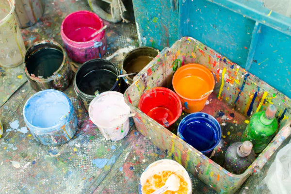 A photo of a variety of different colors of open cans of paint on the floor. Paint has spilled onto the floor producing an almost rainbow like effect.