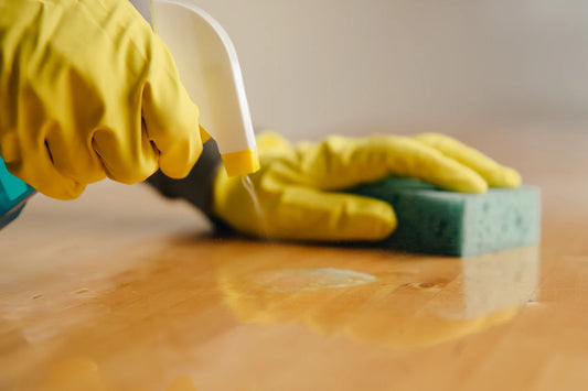 Closeup of human hands. In the right hand is a spray bottle spraying a cleaning agent onto a wooden table. In the left hand is a sponge.